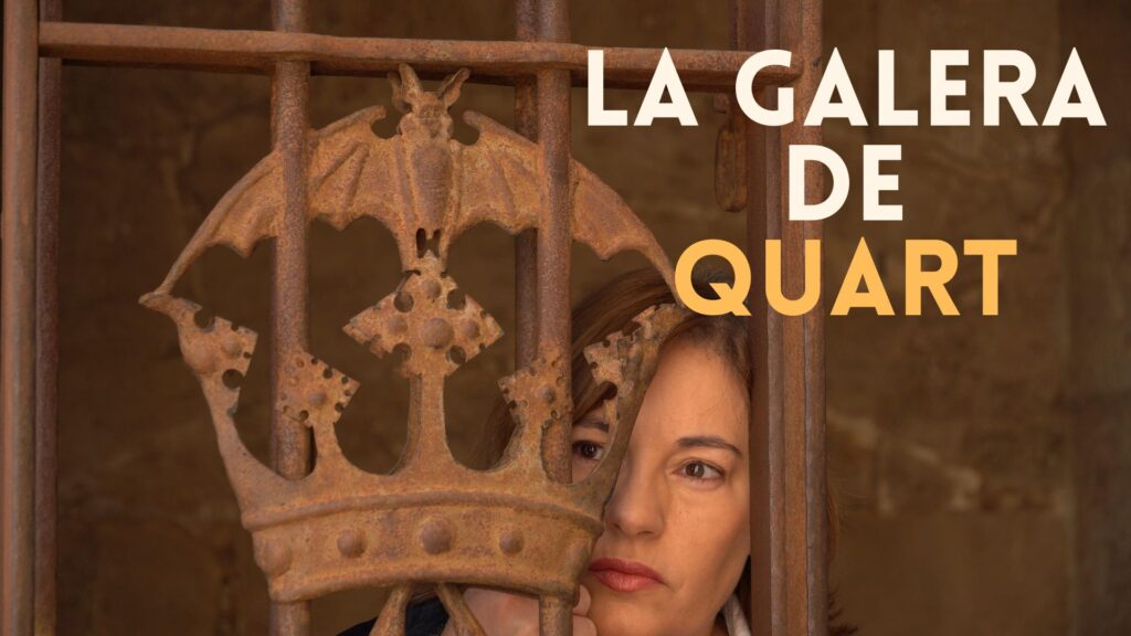 Se muestra parte de la reja de una de las puertas interiores de las Torres de Quart, que representa el escudo de la ciudad (la corona con el murciélago), tras la reja el rostro de una mujer recordando que en lugar se encerraba a mujeres en el pasado.