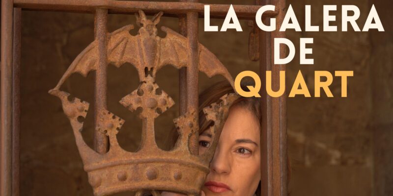 Se muestra parte de la reja de una de las puertas interiores de las Torres de Quart, que representa el escudo de la ciudad (la corona con el murciélago), tras la reja el rostro de una mujer recordando que en lugar se encerraba a mujeres en el pasado.
