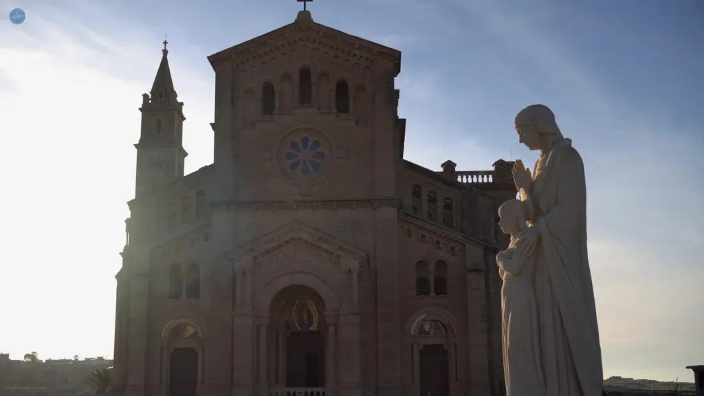 Fachada neogótica de la Basílica del Santuario de Ta' Pinu en Gozo, con la estatua de la Virgen en primer plano al contraluz del atardecer