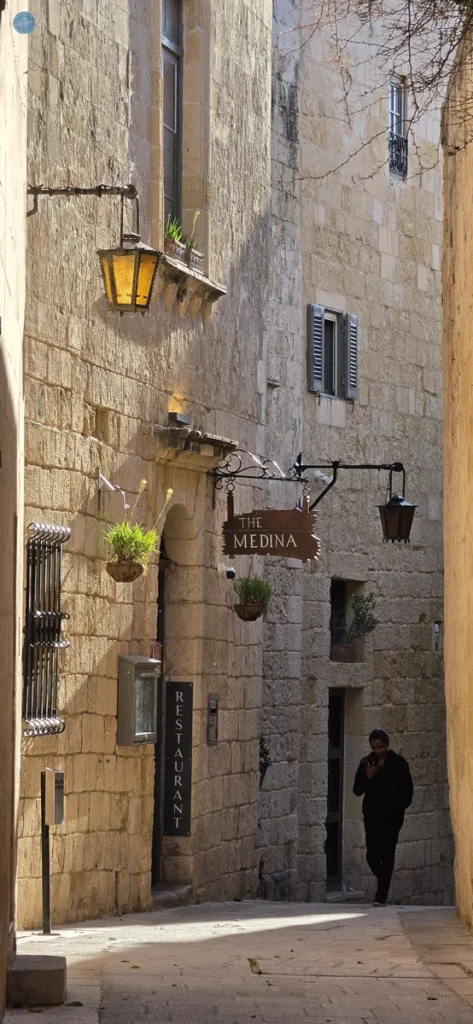 Callejuela de piedra caliza en Mdina, la ciudad medieval amurallada de Malta, con faroles y el cartel del restaurante The Medina